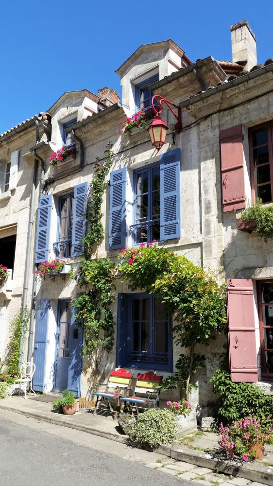 Brantome house White painted 3 story house with blue shutters and greenery on walls , 2 coloured wooden seats rest against the outside