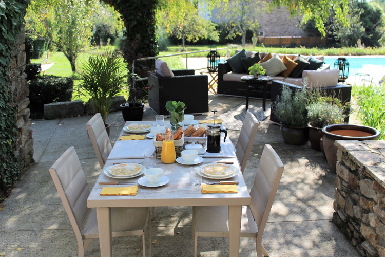 A beige table and four chairs on a patio set out for breakfast with orange juice and croissants
