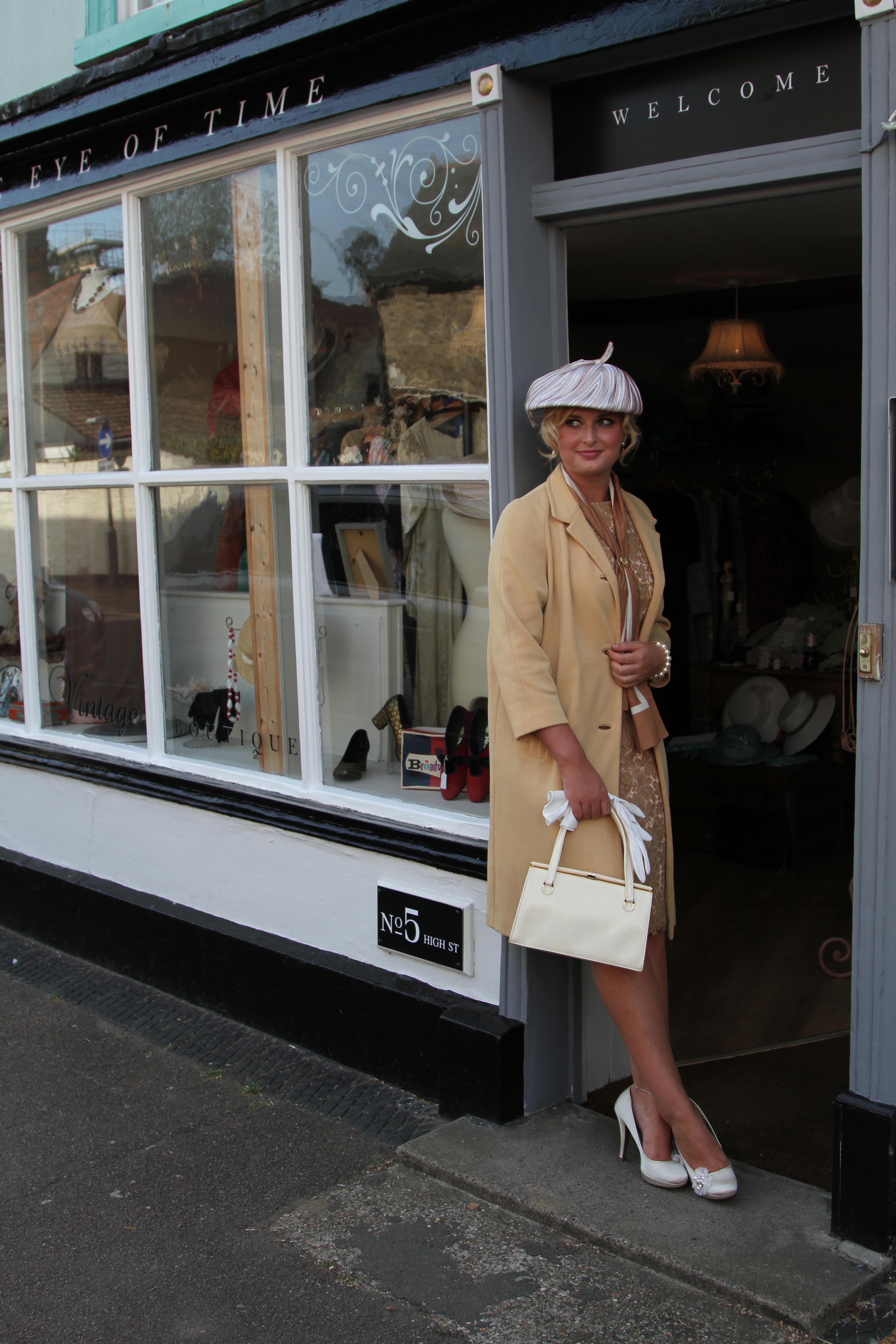 Girl with vintage beige coat, white hat, gloves, bag and shoes leaning against a shop doorwayv